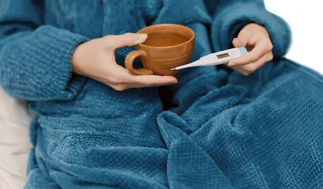 Person in blue blanket holding thermometer and cup of coffee on bed two young men studying together